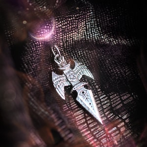 May include: A silver pendant featuring a stylized dagger with wings. The dagger has intricate details and a small eye symbol at the top. The pendant is hanging on a black textured surface.