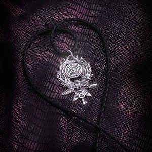 May include: A silver pendant with a detailed design of a bird with outstretched wings, a key, and a circle with a symbol inside. The pendant is hanging on a black leather cord.