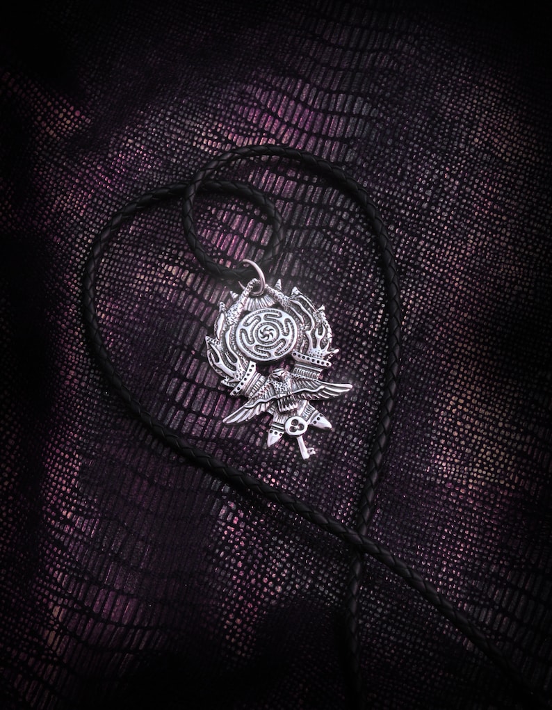 May include: A silver pendant with a detailed design of a bird with outstretched wings, a key, and a circle with a symbol inside. The pendant is hanging on a black leather cord.