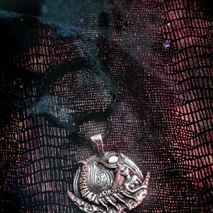 May include: Silver pendant necklace with a detailed design of a woman holding a scorpion. The pendant is on a black leather cord and is set against a dark red background.