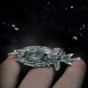 May include: A silver pendant featuring a detailed design of a bird with intricate wings and a spiral pattern. The pendant is held in a hand.
