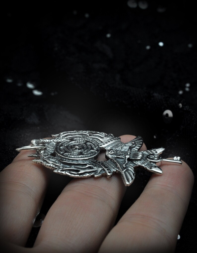 May include: A silver pendant featuring a detailed design of a bird with intricate wings and a spiral pattern. The pendant is held in a hand.