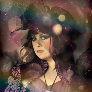May include: A woman wearing a black lace veil and a purple dress with a silver pendant necklace. The background is blurred and has a soft, dreamy effect.