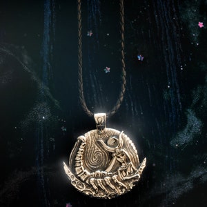May include: A gold pendant necklace with a detailed design of a woman holding a crescent moon and a scorpion. The pendant is on a black leather cord.