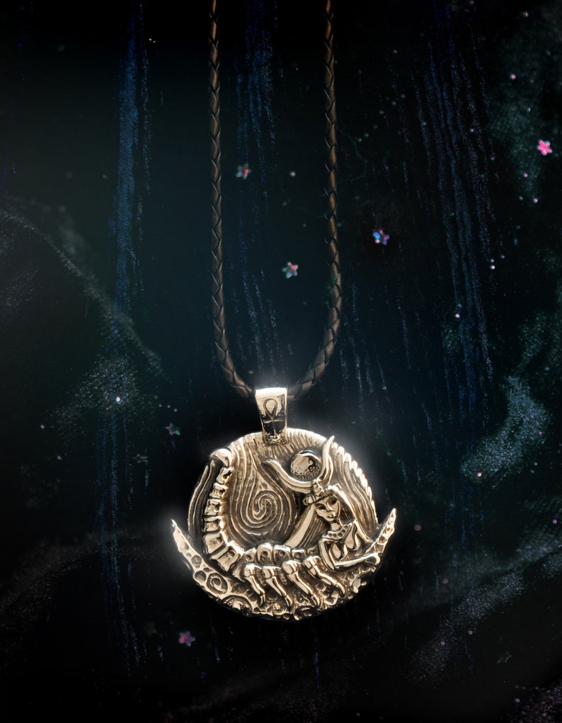 May include: A gold pendant necklace with a detailed design of a woman holding a crescent moon and a scorpion. The pendant is on a black leather cord.