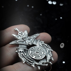 May include: A silver pendant featuring a detailed design of a winged creature holding a circular symbol with a spiral pattern. The pendant has a loop for attaching a chain.