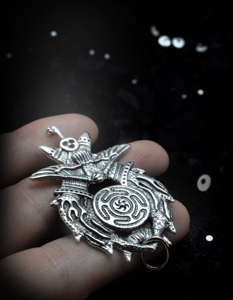 May include: A silver pendant featuring a detailed design of a winged creature holding a circular symbol with a spiral pattern. The pendant has a loop for attaching a chain.
