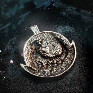 May include: A silver pendant with a textured surface and a small heart with the inscription "AMMM 925" in the center.