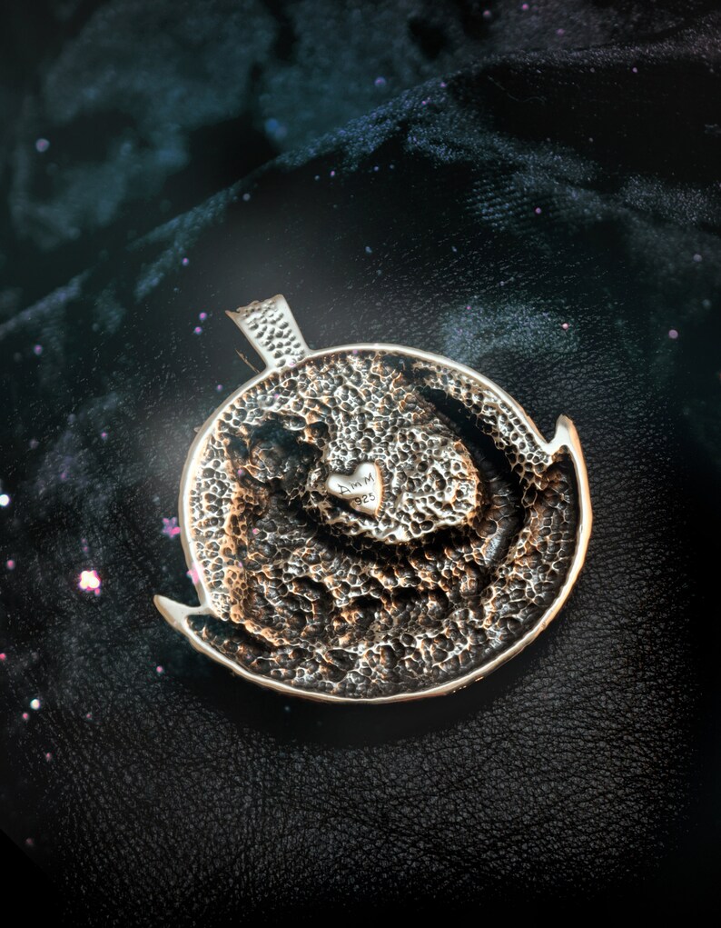 May include: A silver pendant with a textured surface and a small heart with the inscription "AMMM 925" in the center.