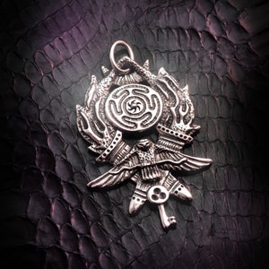 May include: A silver pendant featuring a stylized bird with outstretched wings, a circular design with a spiral pattern, and a key-like detail. The pendant is on a dark textured background.