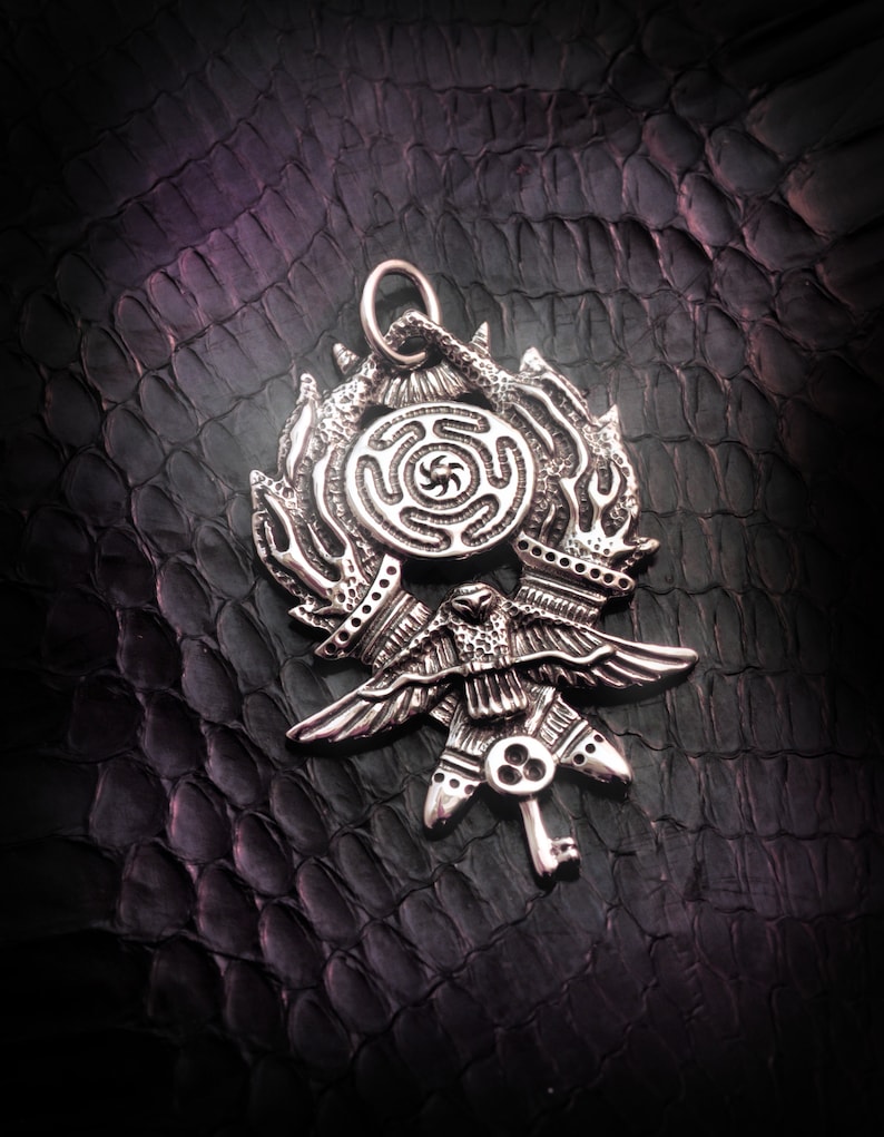 May include: A silver pendant featuring a stylized bird with outstretched wings, a circular design with a spiral pattern, and a key-like detail. The pendant is on a dark textured background.