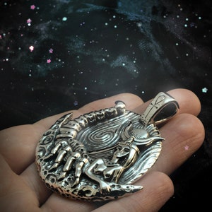 May include: Silver pendant featuring a detailed depiction of a woman with a scorpion and a crescent moon. The pendant has a textured surface and a loop for attaching a chain.