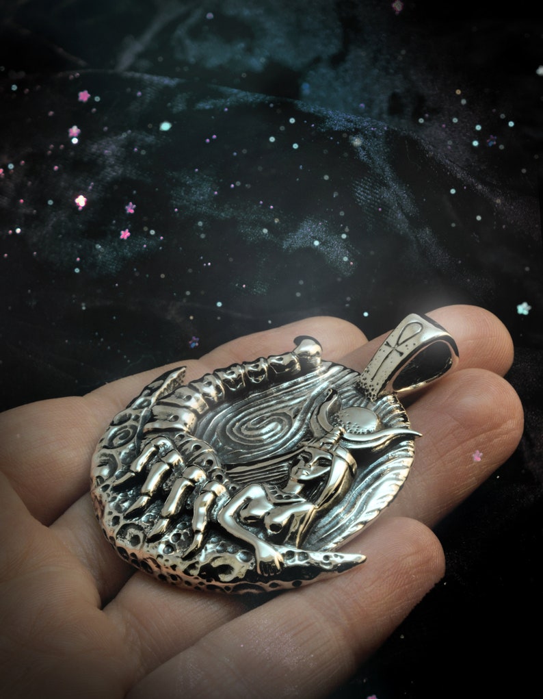 May include: Silver pendant featuring a detailed depiction of a woman with a scorpion and a crescent moon. The pendant has a textured surface and a loop for attaching a chain.