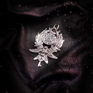 May include: A silver pendant featuring a stylized bird with outstretched wings, a key, and a circular design with a spiral pattern. The pendant is on a dark, textured background.