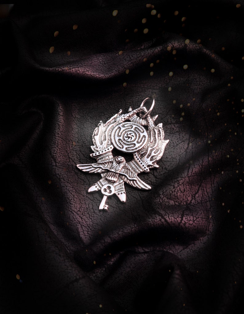 May include: A silver pendant featuring a stylized bird with outstretched wings, a key, and a circular design with a spiral pattern. The pendant is on a dark, textured background.