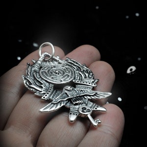 May include: A silver pendant featuring a detailed design of a bird with outstretched wings, a key, and a circular maze pattern.