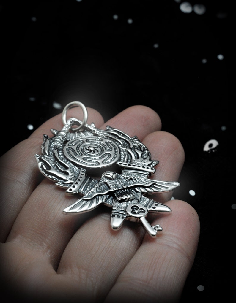 May include: A silver pendant featuring a detailed design of a bird with outstretched wings, a key, and a circular maze pattern.