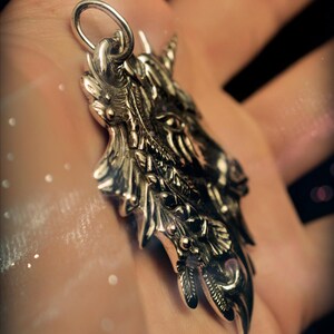May include: A silver pendant featuring a detailed, ornate design of a unicorn's head with a horn, leaves, and flowers. The pendant is on a hand.
