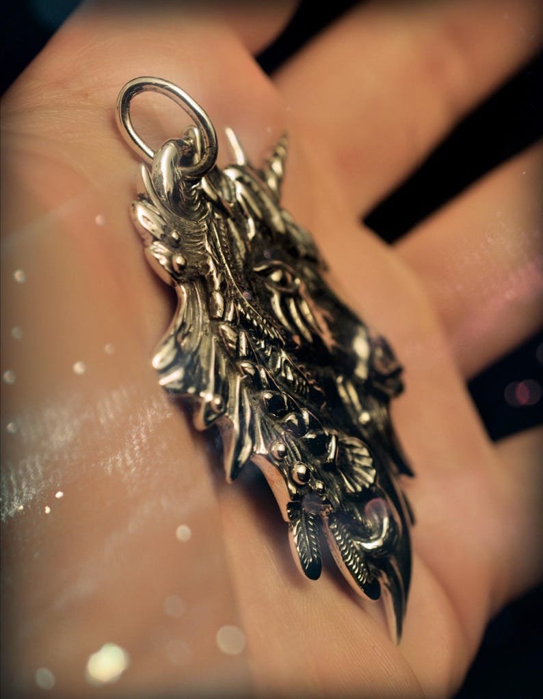 May include: A silver pendant featuring a detailed, ornate design of a unicorn's head with a horn, leaves, and flowers. The pendant is on a hand.