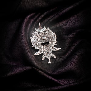 May include: Silver pendant with a detailed design and the word "HEKATE" inscribed on it. The pendant is on a dark fabric background.