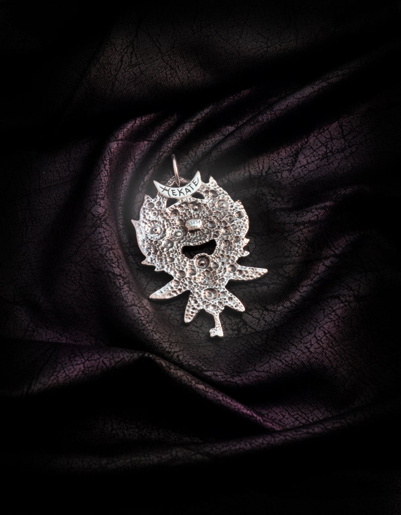May include: Silver pendant with a detailed design and the word "HEKATE" inscribed on it. The pendant is on a dark fabric background.