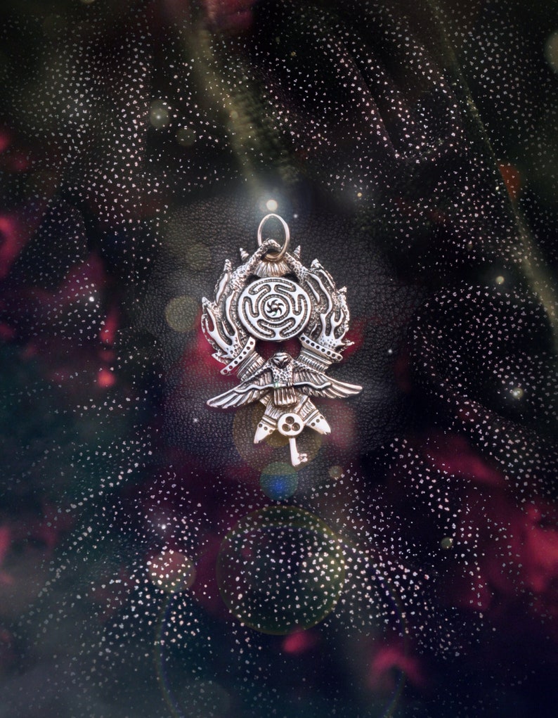 May include: A silver pendant with a keyhole design and a circular symbol with a swastika-like pattern. The pendant is surrounded by a ring of stylized hands. The pendant is against a dark background with a starry pattern.