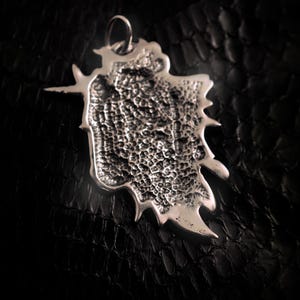 May include: Silver pendant with a textured, organic shape and a small inscription on the back. The pendant has a loop at the top for attaching a chain.