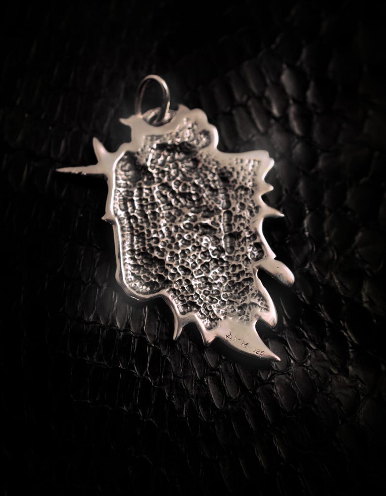 May include: Silver pendant with a textured, organic shape and a small inscription on the back. The pendant has a loop at the top for attaching a chain.