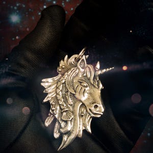 May include: A silver unicorn pendant with intricate details, including a braided mane and a horn. The pendant is set against a dark background.