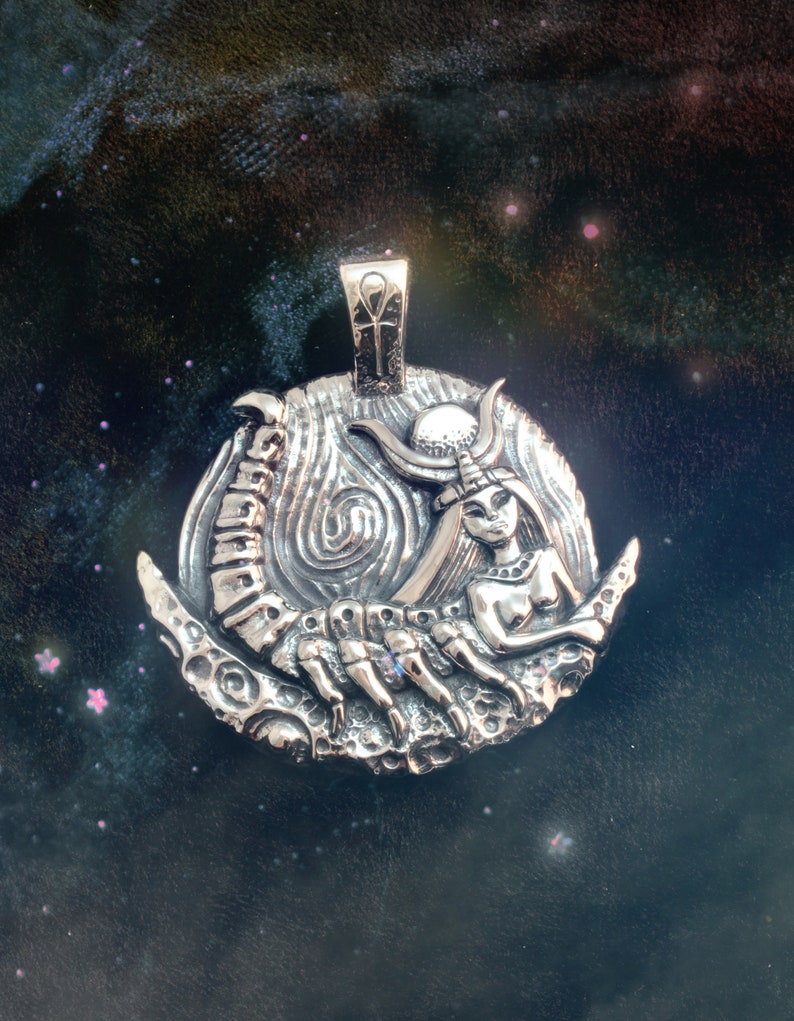 May include: A silver pendant featuring a detailed depiction of a woman with a scorpion tail, set against a crescent moon background. The pendant has an ankh symbol at the top.