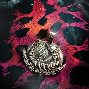 May include: A silver pendant featuring a detailed depiction of a scorpion and a woman with a crown, set against a crescent moon. The pendant is on a pink and black patterned fabric.