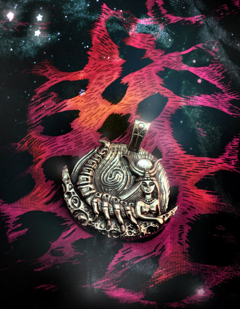 May include: A silver pendant featuring a detailed depiction of a scorpion and a woman with a crown, set against a crescent moon. The pendant is on a pink and black patterned fabric.
