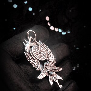 May include: A silver pendant featuring a stylized bird with a key and a circular design with a maze pattern.