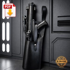 Könnte beinhalten: Ein schwarzes Lederholster für eine Requisiten-Blasterwaffe, bestehend aus einer langen vertikalen Hülle, einem Sicherungsriemen mit silberfarbenem Metalldruckknopf und einem verstellbaren Schulterriemen aus glattem, mattem schwarzem Leder.