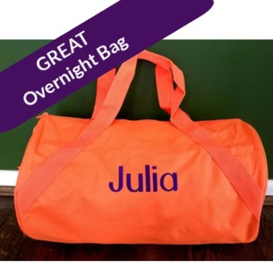May include: A bright orange duffel bag featuring two matching fabric handles and the name Julia printed in purple on the front. A purple diagonal banner across the top left corner displays the white text GREAT Overnight Bag.