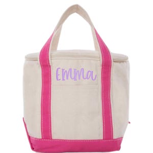 May include: A canvas tote bag with a cream-colored body, accented with bright pink trim and handles. The name "Emma" is embroidered in lavender script on the front. The bag has a zippered top and a rectangular shape.