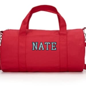 May include: A bright red cylindrical duffel bag featuring two top handles and an adjustable shoulder strap. The name NATE is embroidered in light blue block letters with a dark outline on the center front of the bag, which has a zipper closure.