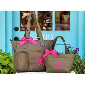 May include: Two olive green tote bags with pink bows and monogrammed with the letters "SH" in pink. The larger bag has a zippered top and multiple pockets. The smaller bag has a top handle and a zippered closure.