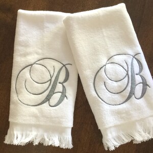 May include: Two white hand towels featuring a decorative gray embroidered letter B in a cursive script font. Each towel has a soft, plush texture and a fringed edge at the bottom, providing a clean and simple look for bathroom or kitchen use.
