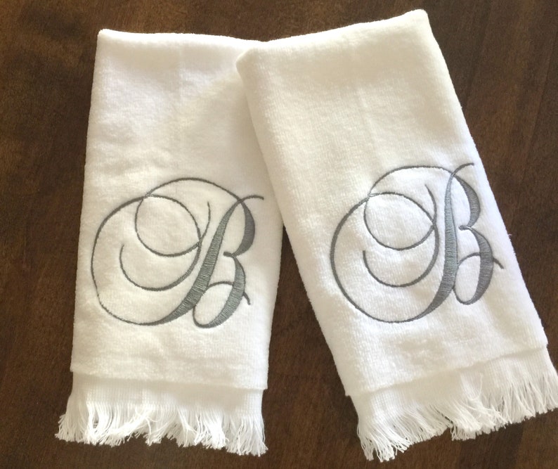 May include: Two white hand towels featuring a decorative gray embroidered letter B in a cursive script font. Each towel has a soft, plush texture and a fringed edge at the bottom, providing a clean and simple look for bathroom or kitchen use.