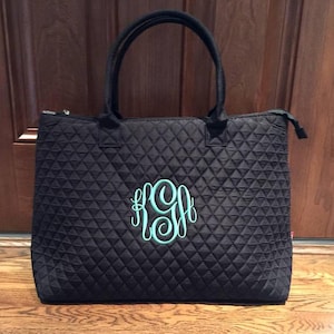 May include: Black quilted tote bag with a teal monogram. The bag has two black handles and a zipper closure. The bag is sitting on a wooden floor. The monogram is in a script font.