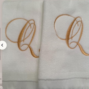 May include: Two white terry cloth towels featuring a large, embroidered letter Q in a metallic gold thread. The letter is rendered in a cursive, calligraphic script style with elegant loops and flourishes centered on each towel fabric surface.