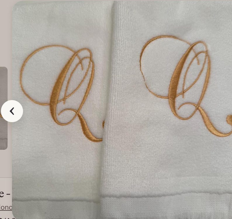 May include: Two white terry cloth towels featuring a large, embroidered letter Q in a metallic gold thread. The letter is rendered in a cursive, calligraphic script style with elegant loops and flourishes centered on each towel fabric surface.