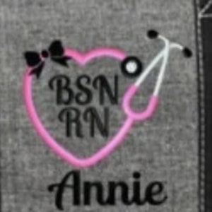 May include: A gray fabric with a pink heart-shaped stethoscope design. The design includes the text "BSN RN" and the name "Annie" in black lettering, along with a black bow.
