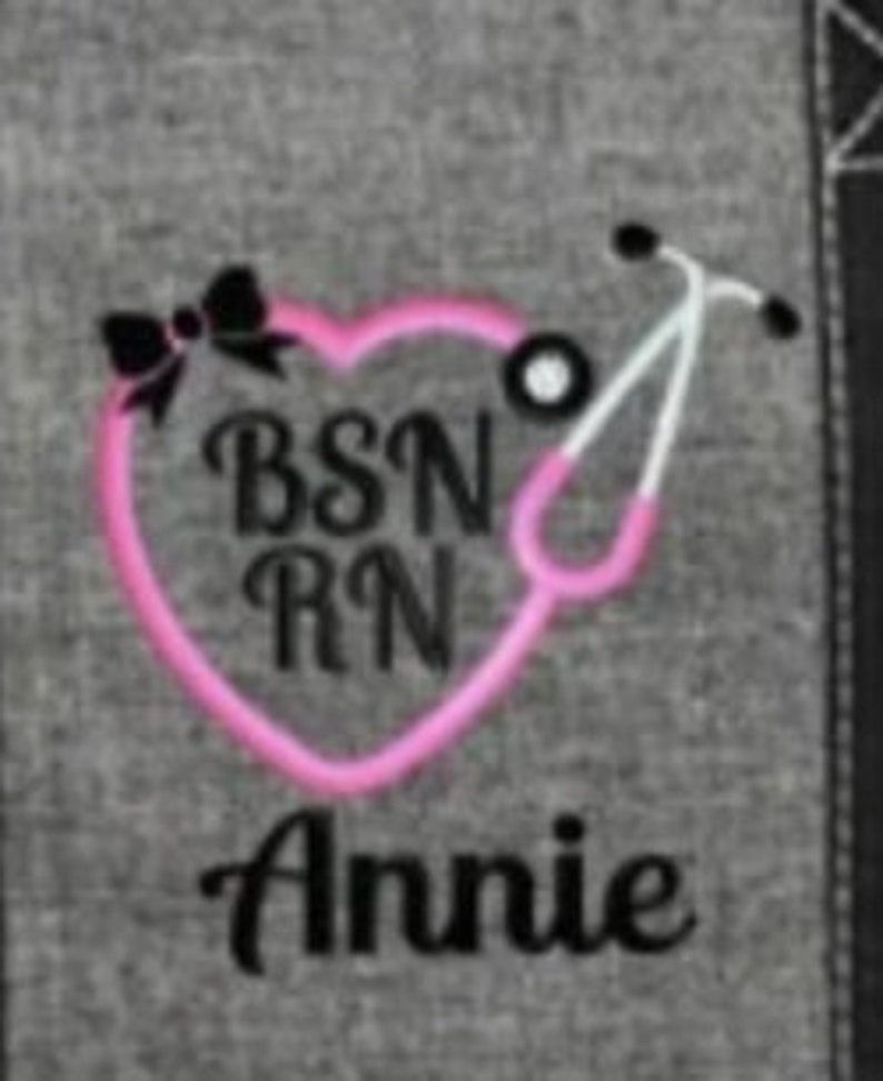 May include: A gray fabric with a pink heart-shaped stethoscope design. The design includes the text "BSN RN" and the name "Annie" in black lettering, along with a black bow.