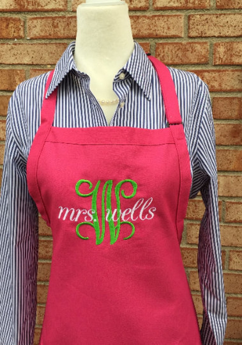 Pu&ograve; includere: Un grembiule rosa con spalline regolabili e un monogramma ricamato in verde che dice "mrs. wells"