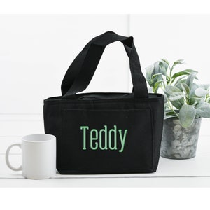 May include: A black rectangular lunch bag with two black fabric handles and a zipper closure. The front features the name Teddy embroidered in light green capital letters. The bag has a flat base and a structured design for carrying items.