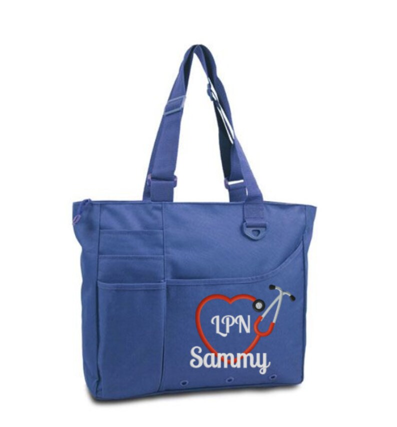 Pode incluir: Uma bolsa tote azul com um bolso grande na frente. A bolsa tem um cora&ccedil;&atilde;o vermelho com um estetosc&oacute;pio e o texto "LPN Sammy" bordado.