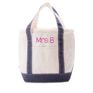 May include: A cream-colored canvas tote bag with navy blue trim and handles. The bag features a zippered top closure and a front exterior pocket. The text Mrs. B is embroidered in pink thread on the front center of the bag above the pocket.