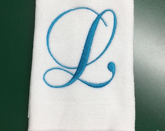 May include: A white rectangular hand towel featuring a textured fabric surface and a decorative fringe along the bottom edge. The center of the towel is embroidered with a large, cursive capital letter L in a vibrant, bright blue thread.